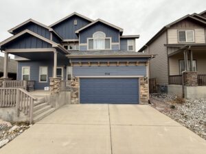 Two-story blue house with a double garage, stone accents, covered front porch, and concrete driveway, set between two similar houses.