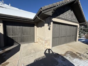 Two closed wooden garage doors on a beige house, with some snow on the roof, ground, and a clear blue sky overhead. A person's shadow is visible in the foreground.