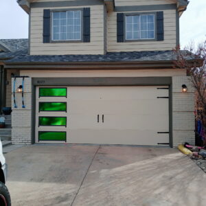 A beige garage door with four rectangular green-tinted windows on the left side, attached to a two-story house with light brick and beige siding.