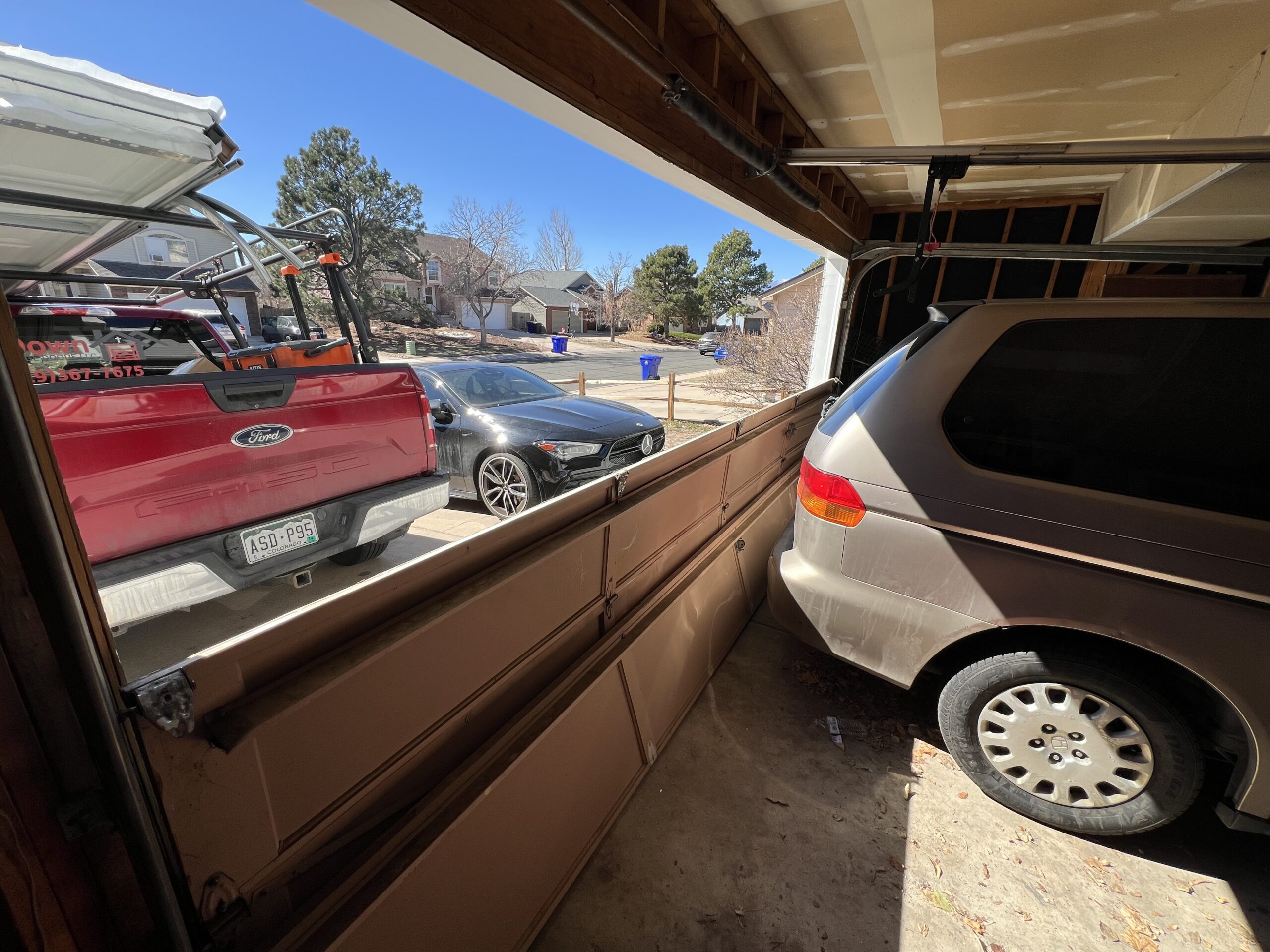 A damaged garage door bent inward rests on a minivan. A red pickup truck and a black car are parked outside on the driveway.