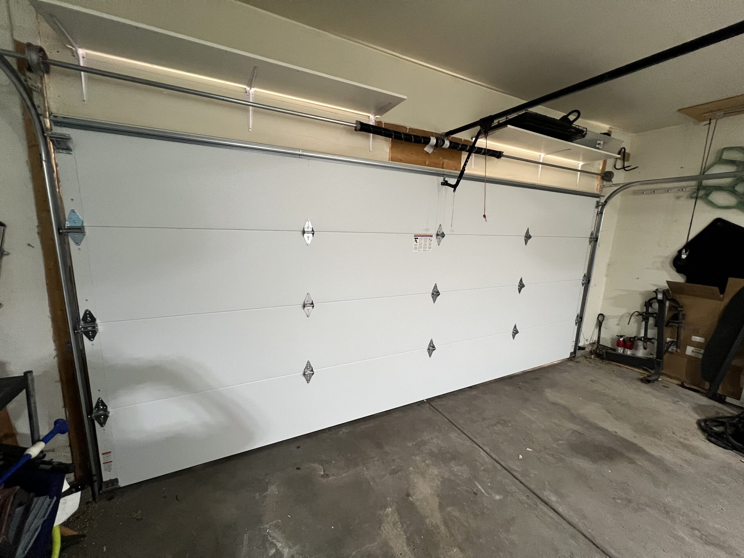 Closed white sectional garage door with metal hinges inside a garage; shelves and assorted items are visible around the space.