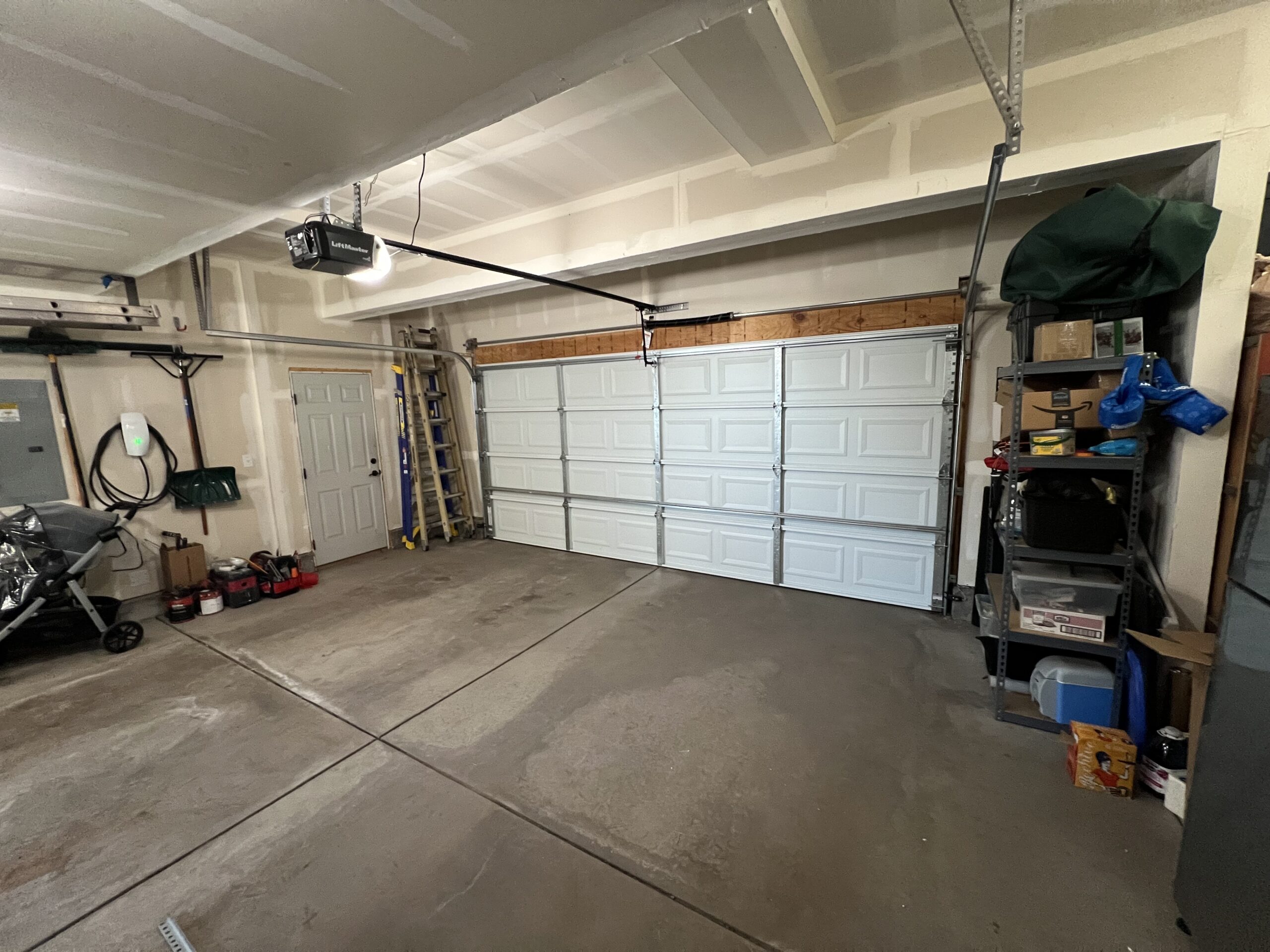A clean, empty garage with a closed double garage door, shelves with assorted items on the right, and tools and ladders stored along the back wall.