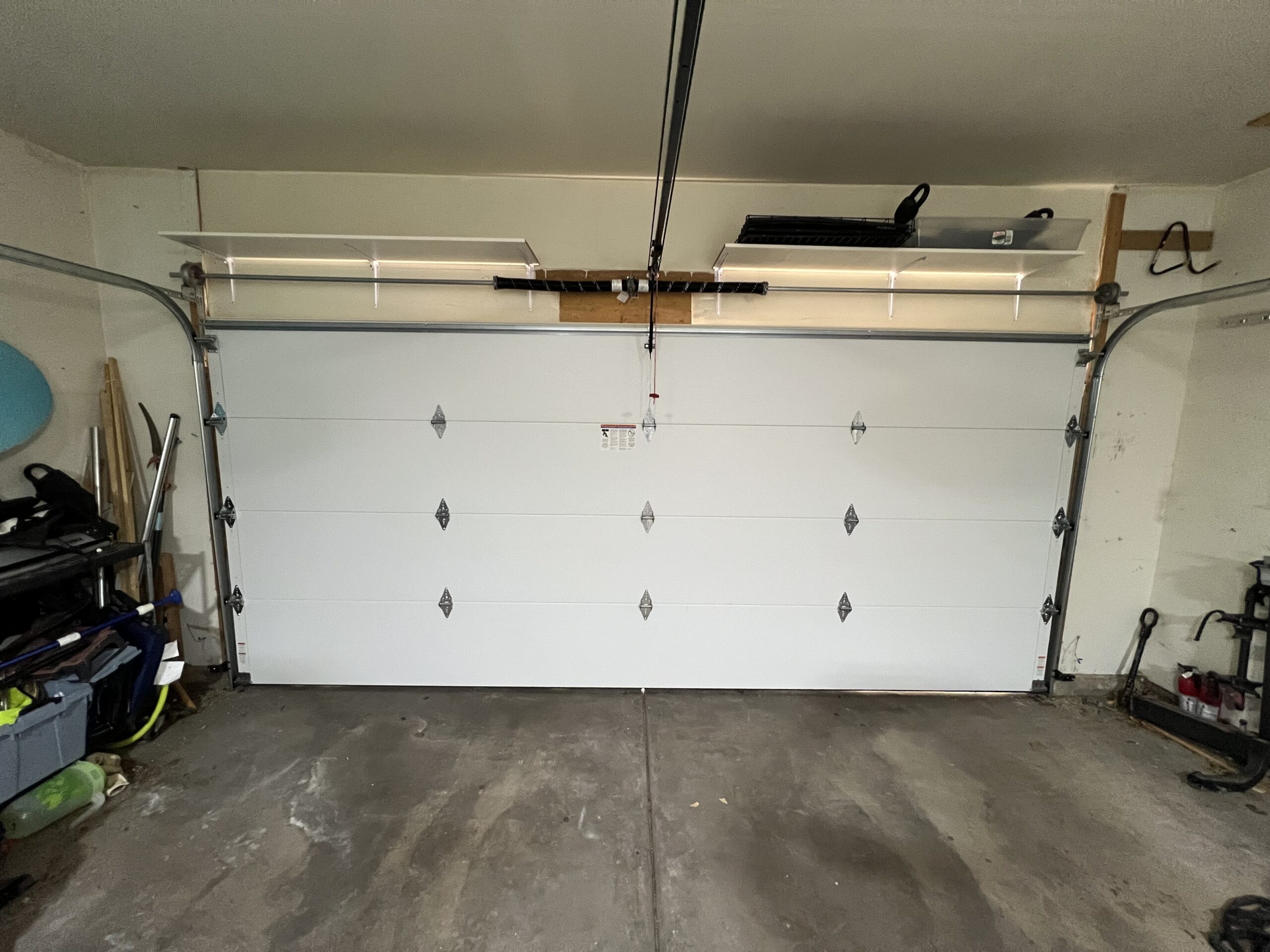 A closed white garage door with four horizontal panels and decorative hardware, viewed from inside a garage with shelving and various items on both sides.