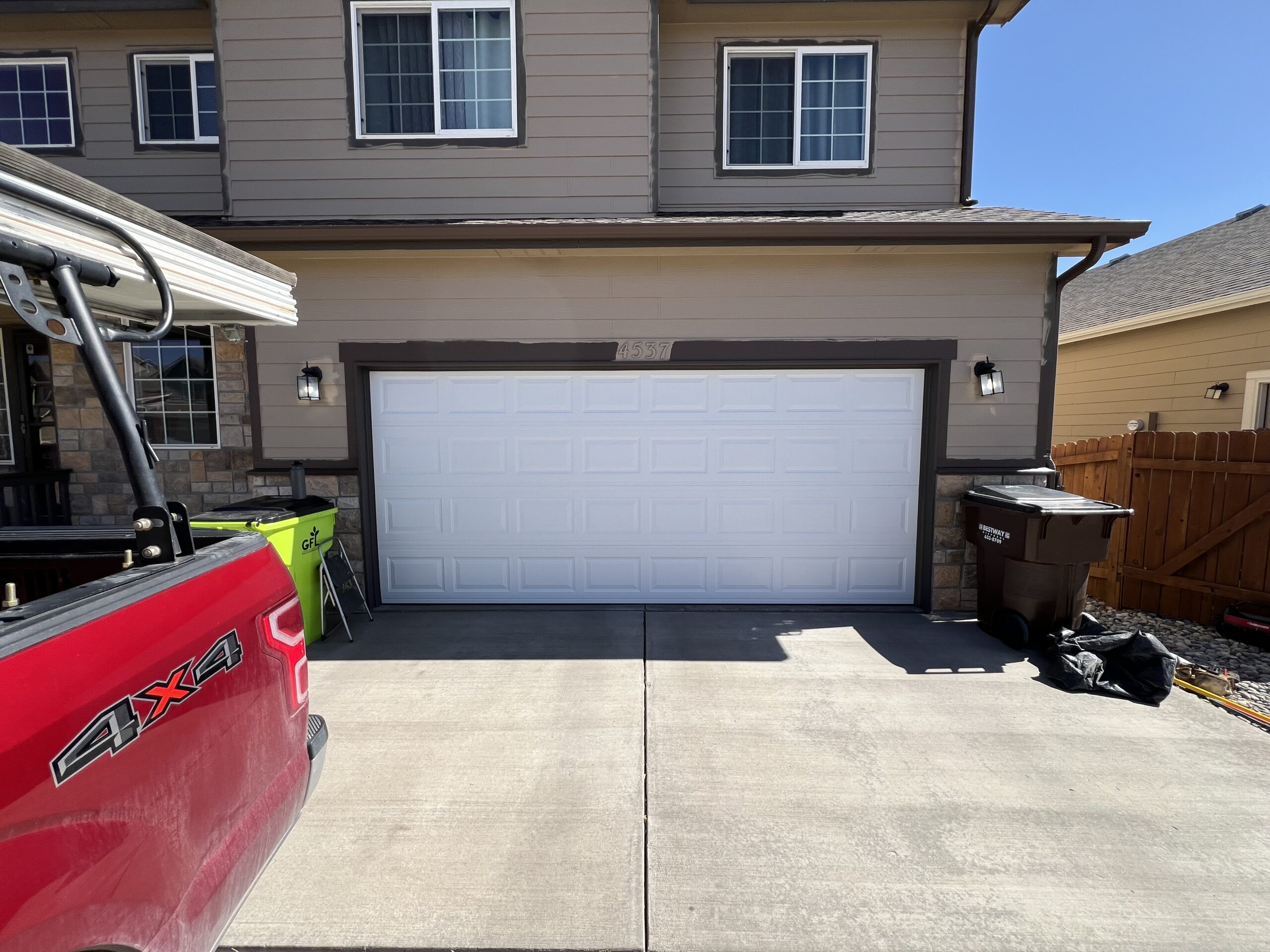 A double garage with a white door is attached to a beige house. A red truck, trash bins, and some items are visible in the driveway and beside the garage.