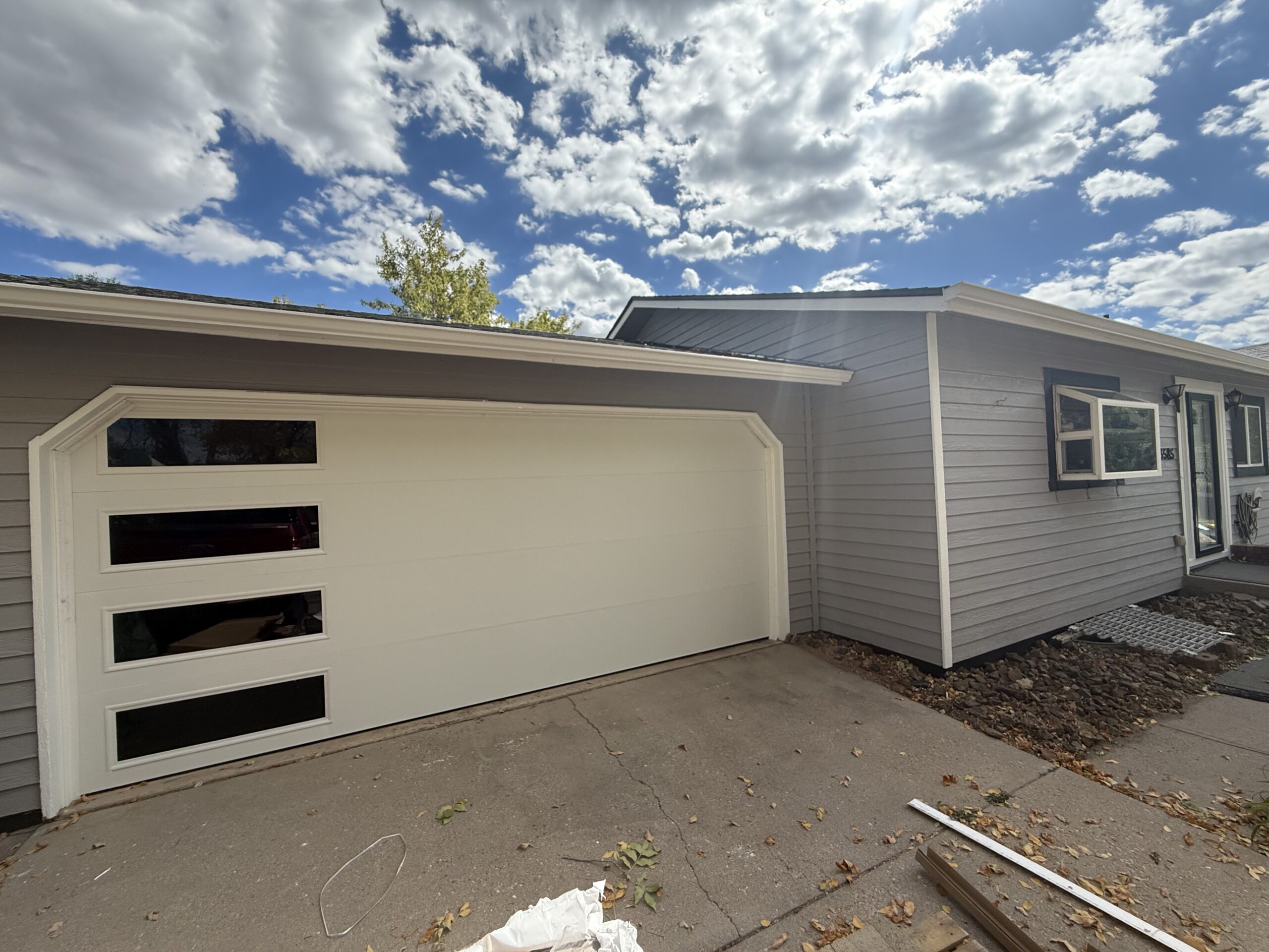 A single-story gray house with a white garage door featuring three horizontal windows, concrete driveway, and partly cloudy sky overhead—showcasing recent garage door repair for added curb appeal and security.