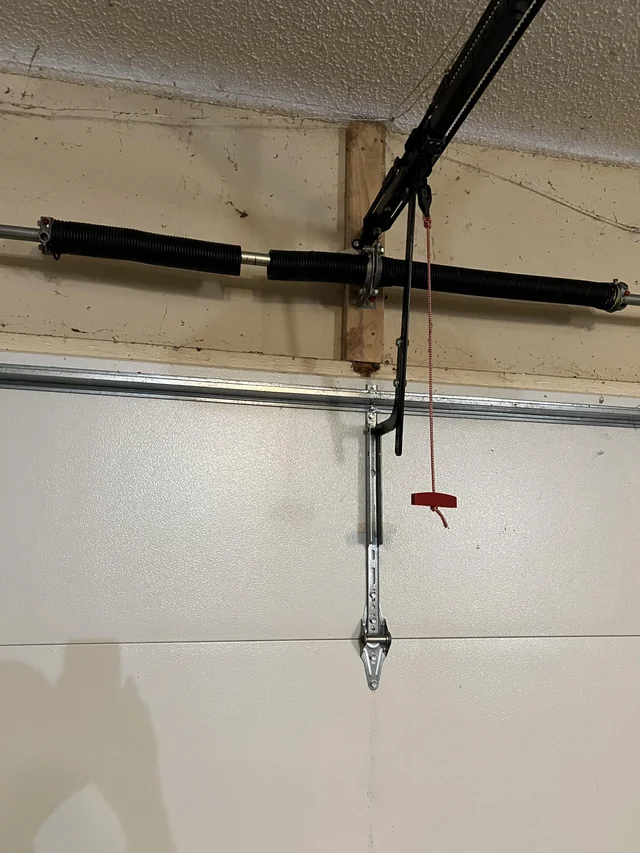 A garage door torsion spring system mounted above a closed white garage door, with a red emergency release cord hanging down.
