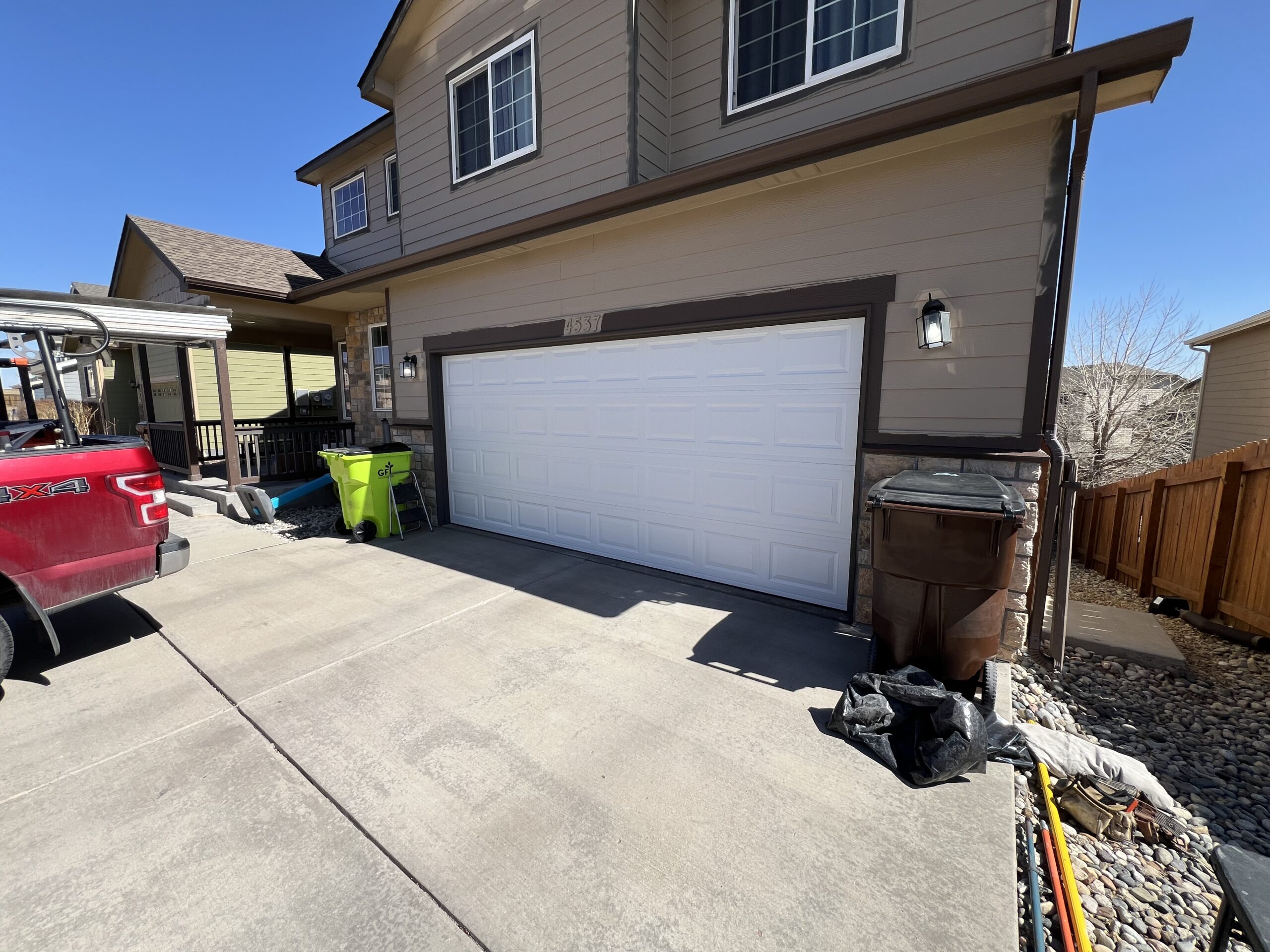 A two-car garage with a closed white door, flanked by trash bins and a red truck parked on the driveway, in front of a tan house on a sunny day.