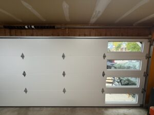 White garage door with four horizontal windows on the right side, viewed from inside a garage with unfinished upper walls and ceiling.